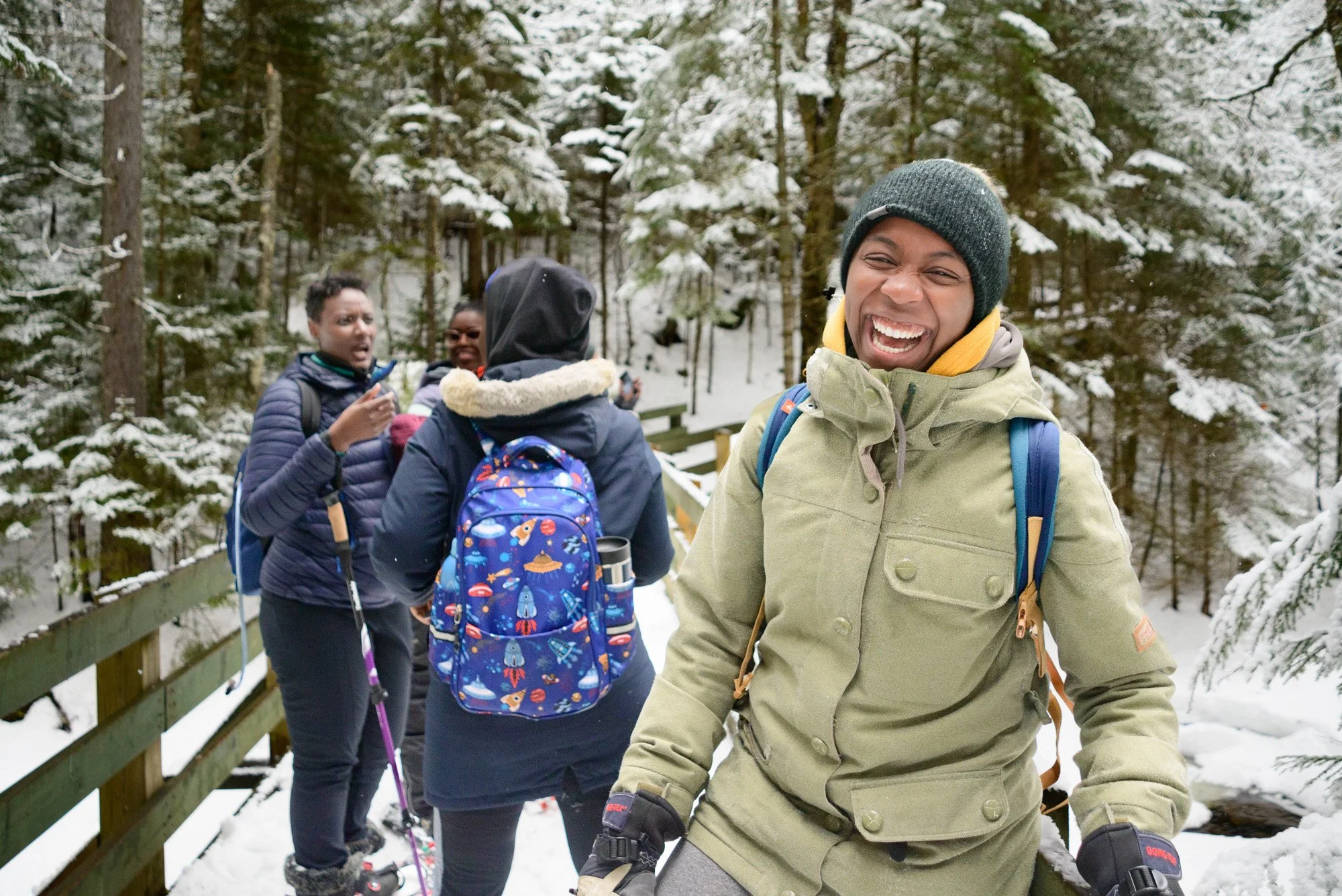 A group of four people enjoying a snowy day outdoors, with one smiling woman in the foreground and three others in the background on a snowy trail surrounded by trees.