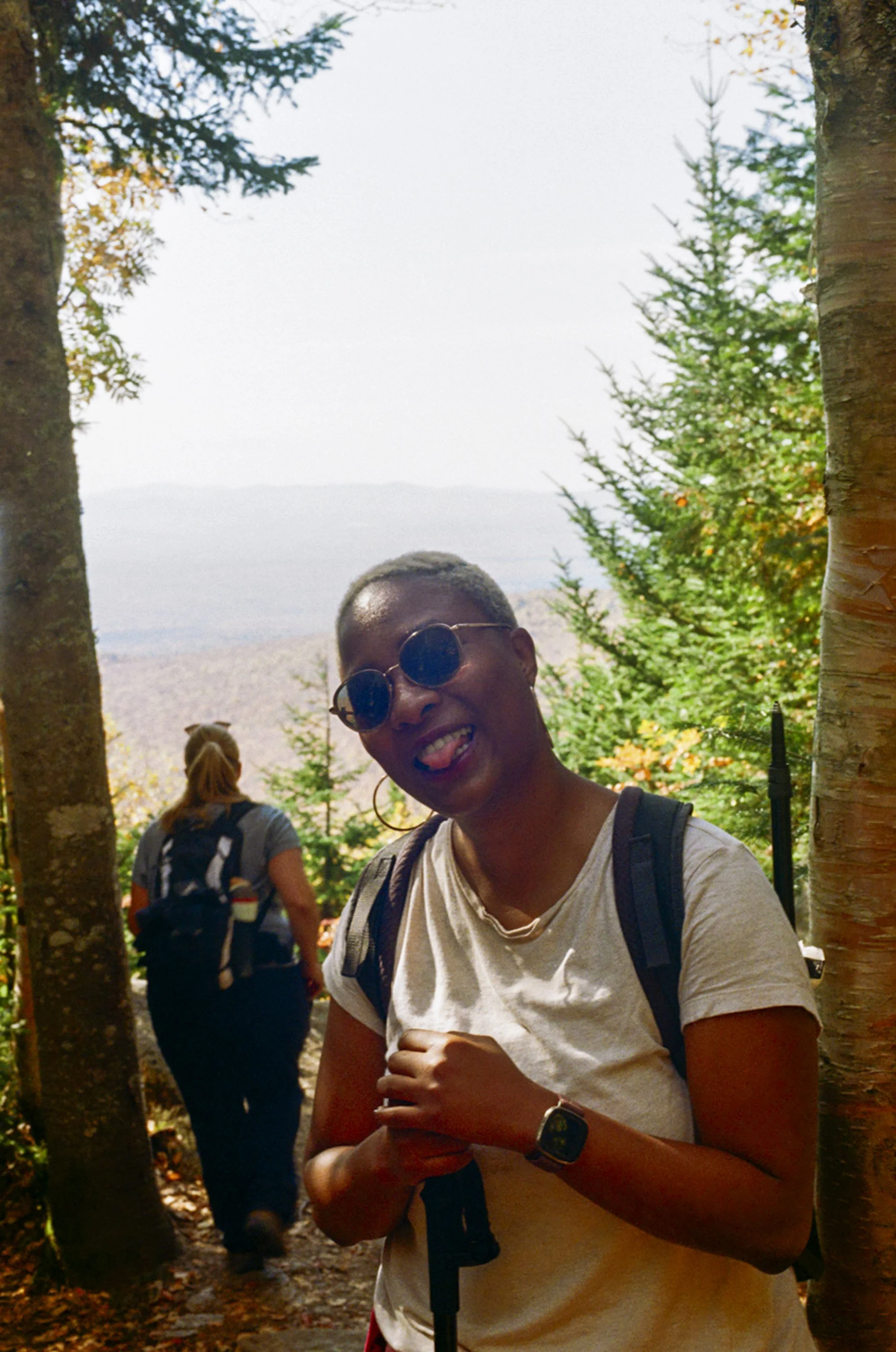 A woman with short hair, wearing sunglasses, a beige t-shirt, a wristwatch, and a backpack, standing outdoors among trees. She is smiling and sticking out her tongue. Other people are in the background.