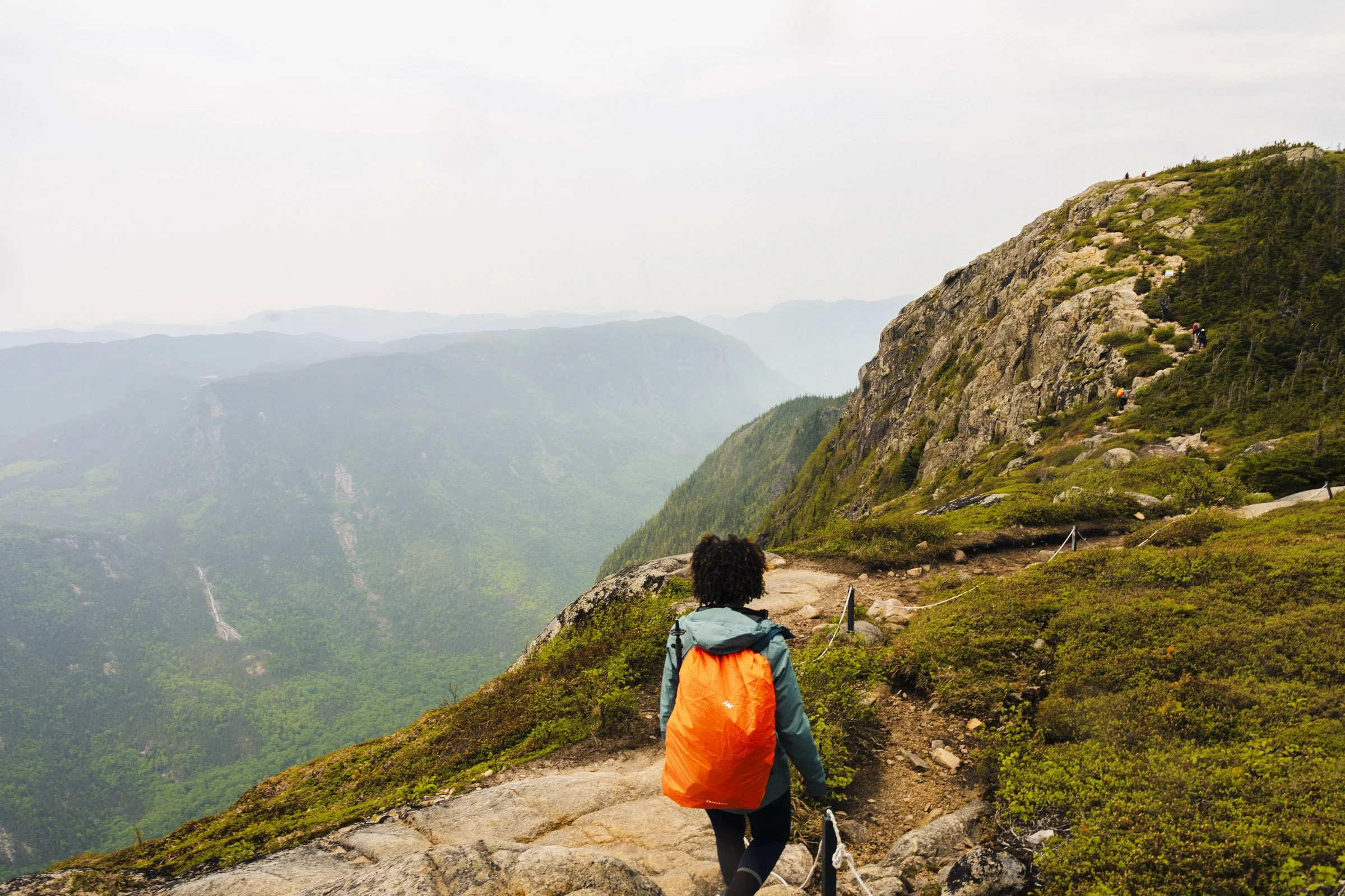 Hikers walking along a mountain trail with a scenic view of mountains and waterfalls in the distance.