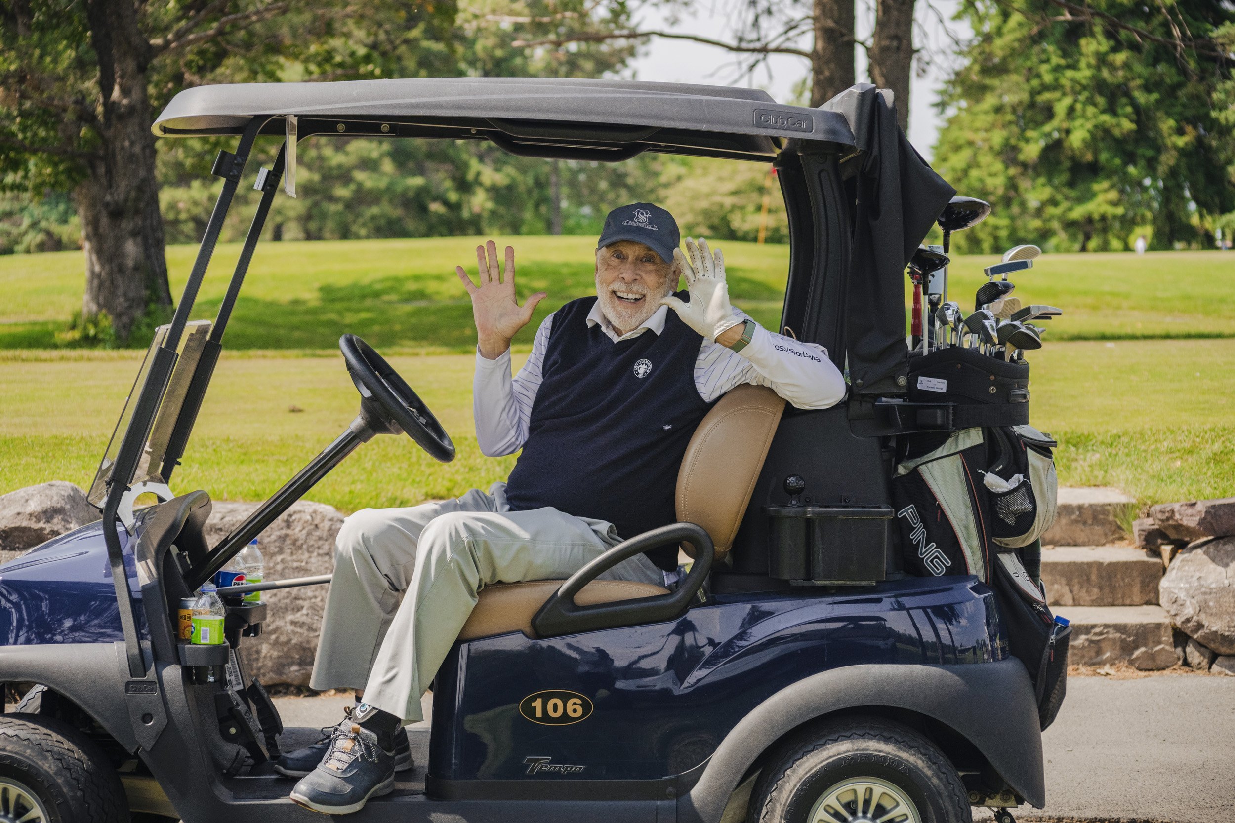 An elderly man sitting in a golf cart on a golf course, smiling and waving, with golf clubs in a bag behind him.