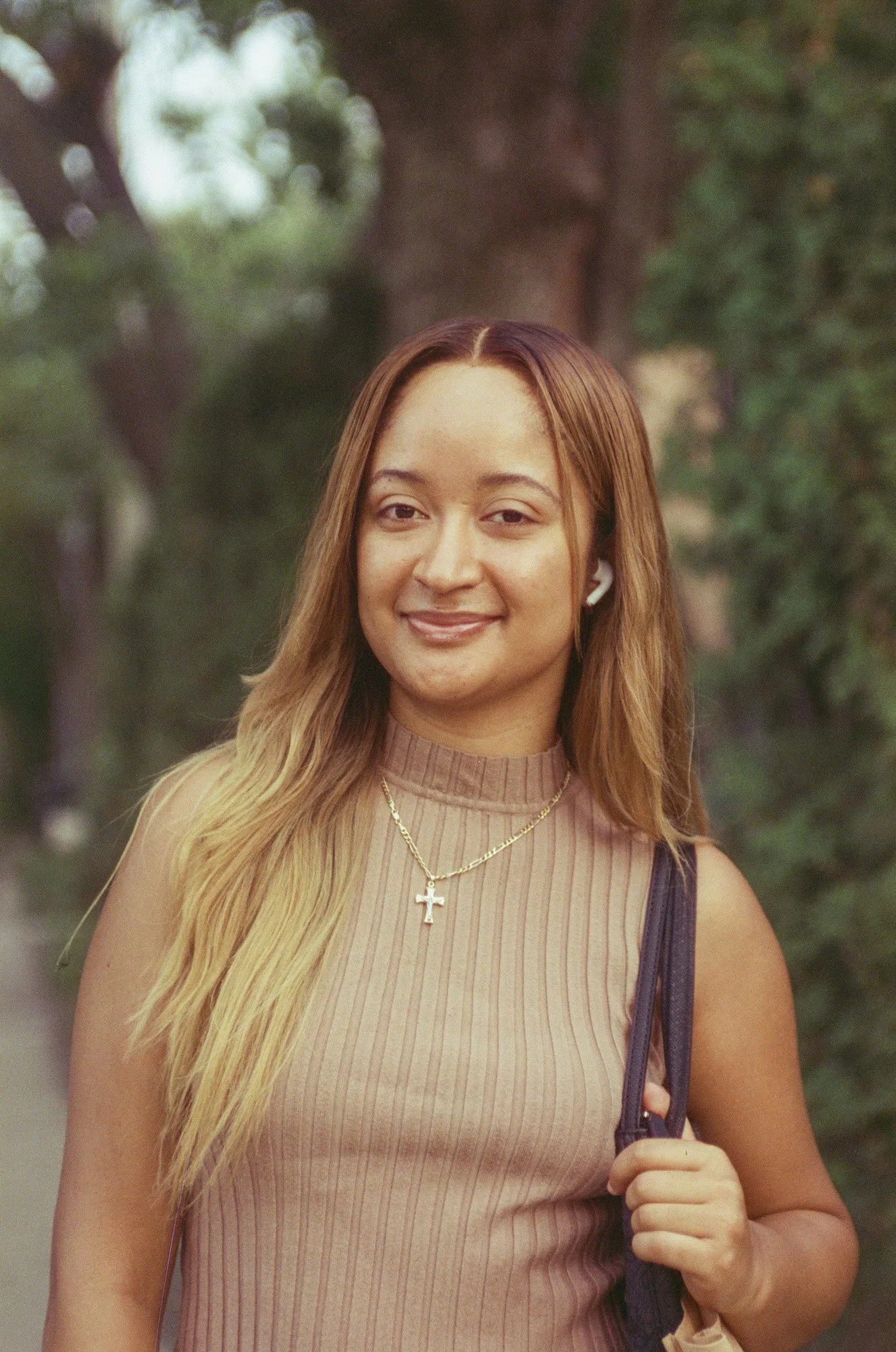 A young woman with long, wavy blonde hair wearing a beige sleeveless ribbed top, a gold necklace with a cross pendant, and white earbuds. She is holding a dark bag strap in her right hand and smiling at the camera, standing outdoors with blurred gree