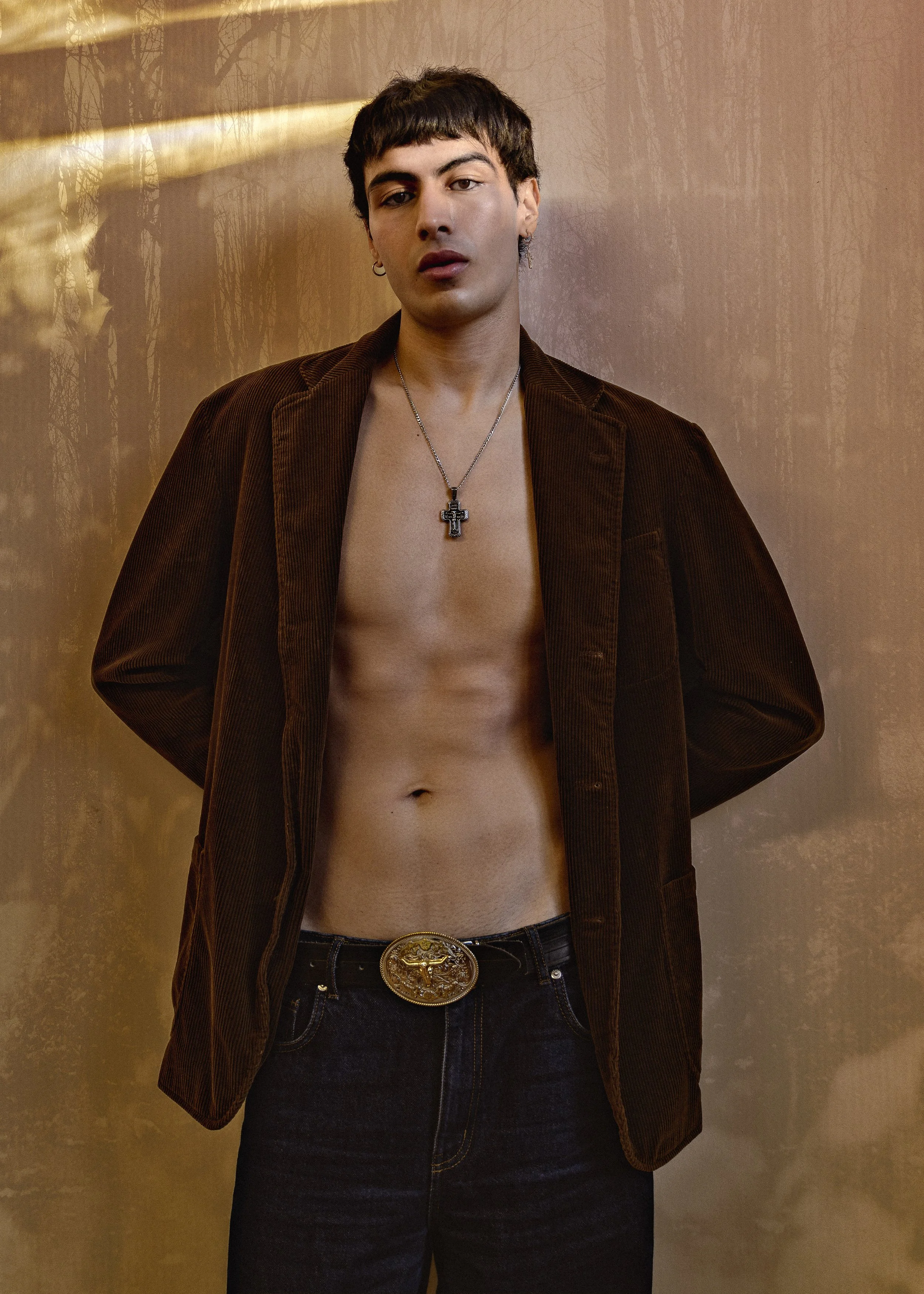 A young man with dark hair and earrings standing shirtless, wearing a brown blazer, black jeans, a large gold belt buckle, and a chain with a cross pendant, against a textured background.