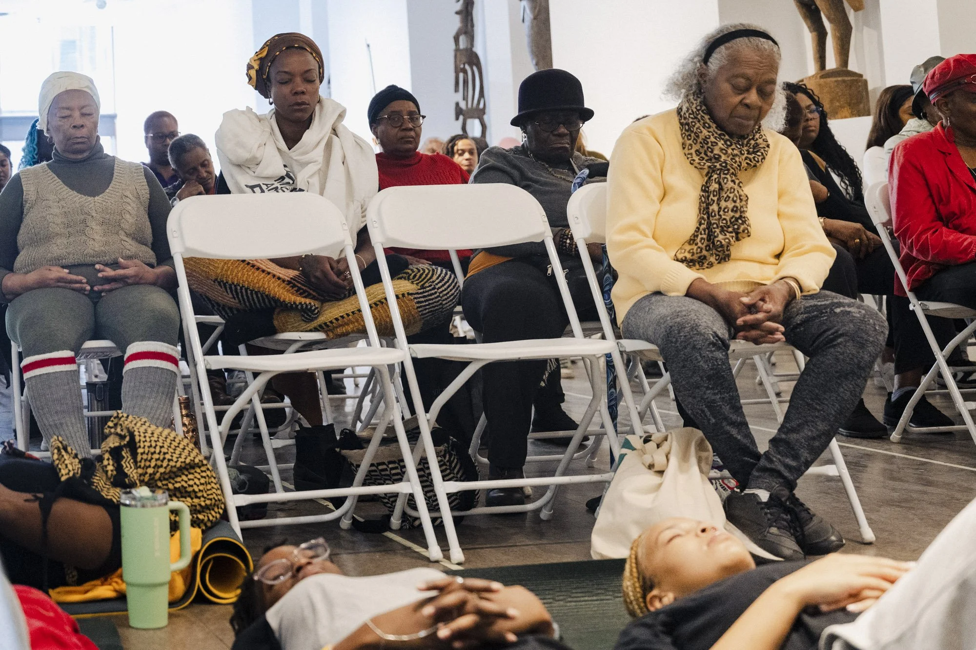 People sitting with eyes closed, participating in a meditation or prayer session in a large room, some with hands on their laps or chest, and various personal belongings and mats on the floor.