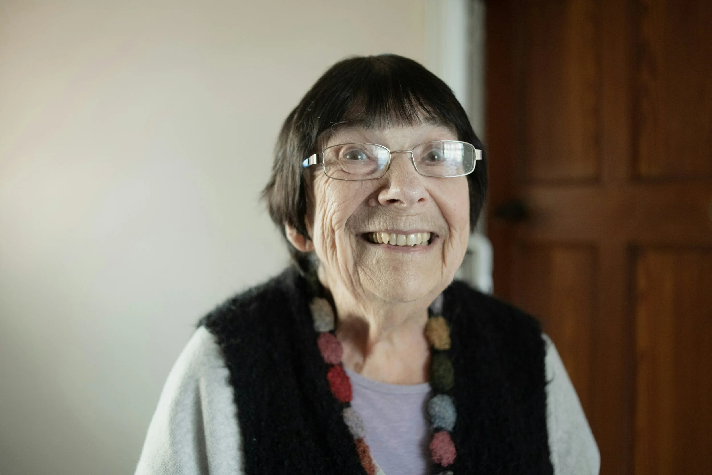 A smiling elderly woman with glasses, short dark hair, wearing a sweater and a colorful scarf, standing indoors near a wooden door.