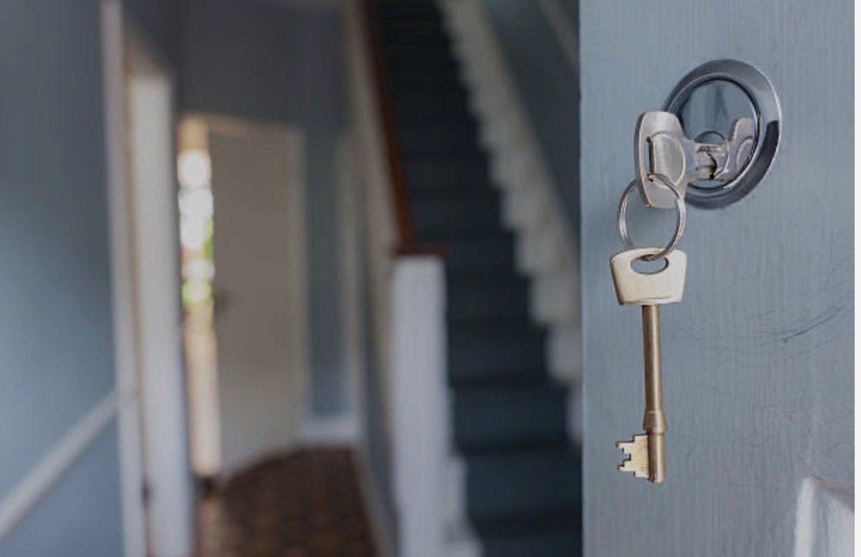 A house key hanging on a key hook on a door, with a staircase visible in the background.
