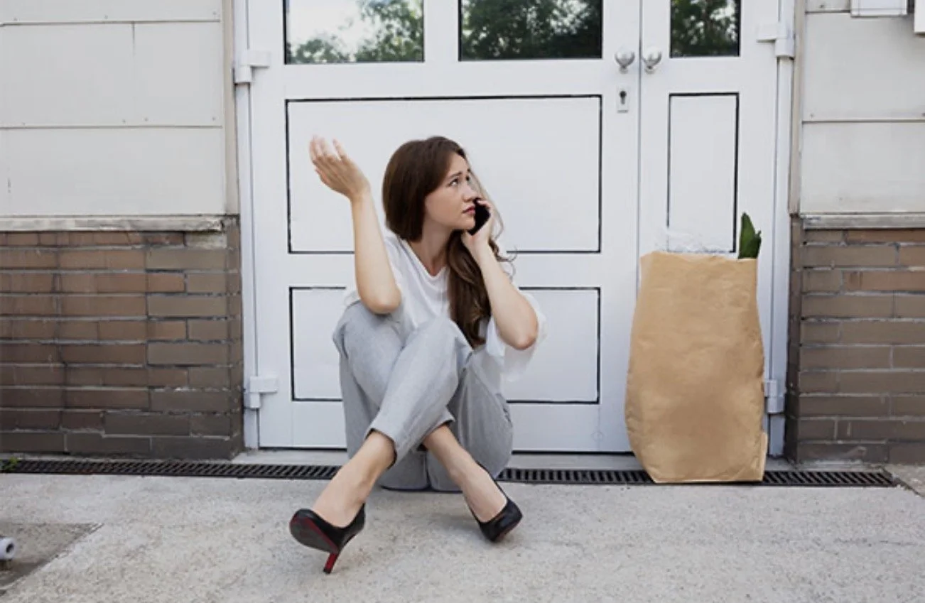 A woman sitting on the ground outside a building, talking on her cellphone, with a neutral expression. She is wearing gray pajamas and black high heels, and there is a large paper bag with a plant in it next to her.