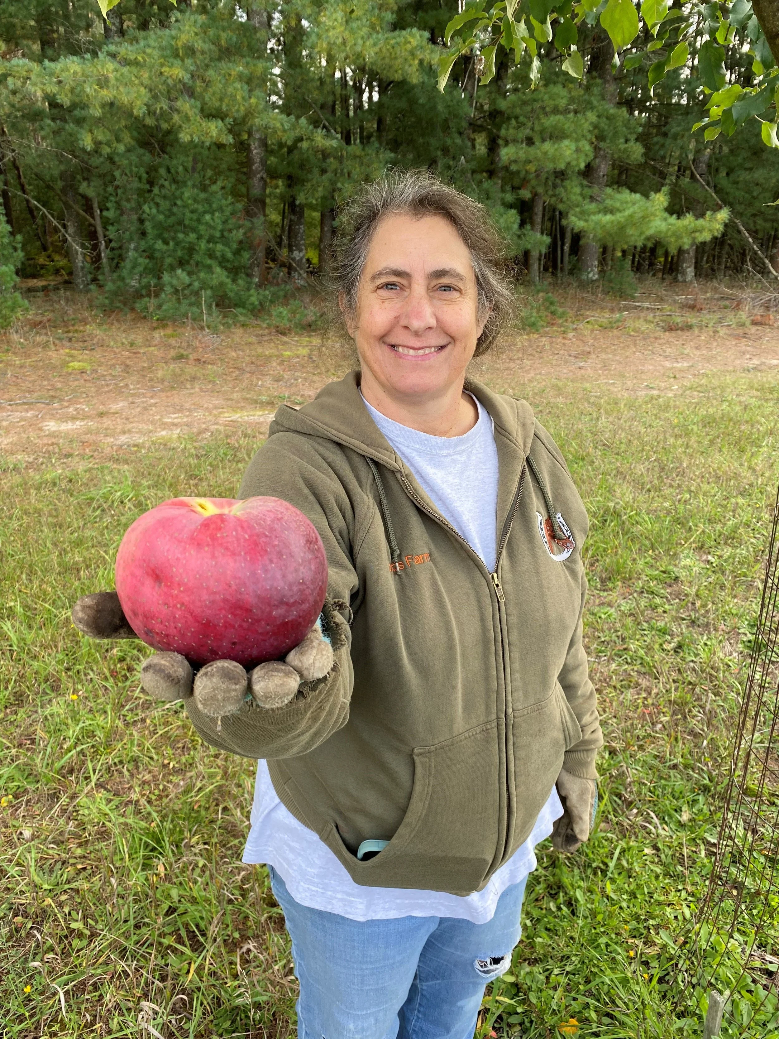 A woman in an olive green hoodie and ripped jeans holding a large, freshly picked red apple with a tree and grassy area in the background.