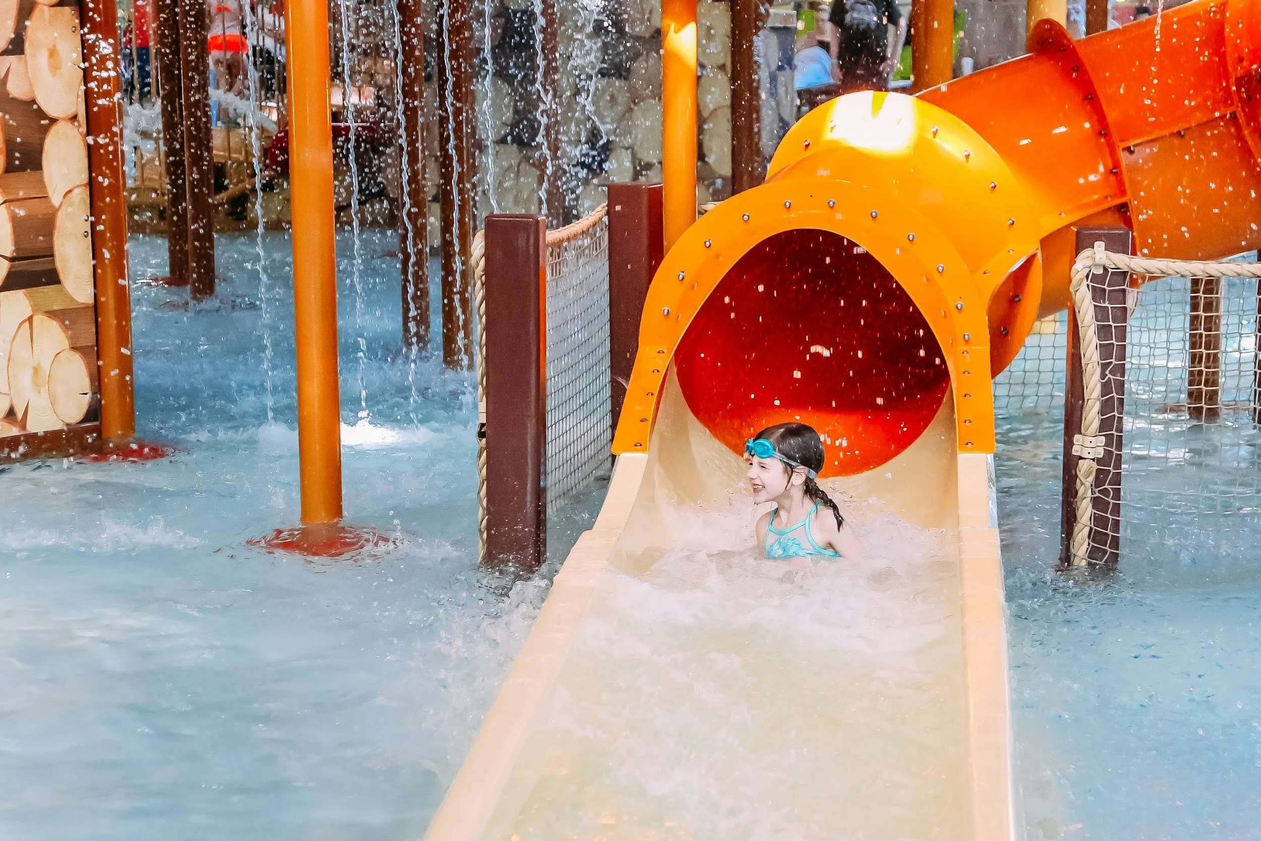 Child playing on water slide at an indoor water park, with water splashing and a bright orange tube-shaped slide.