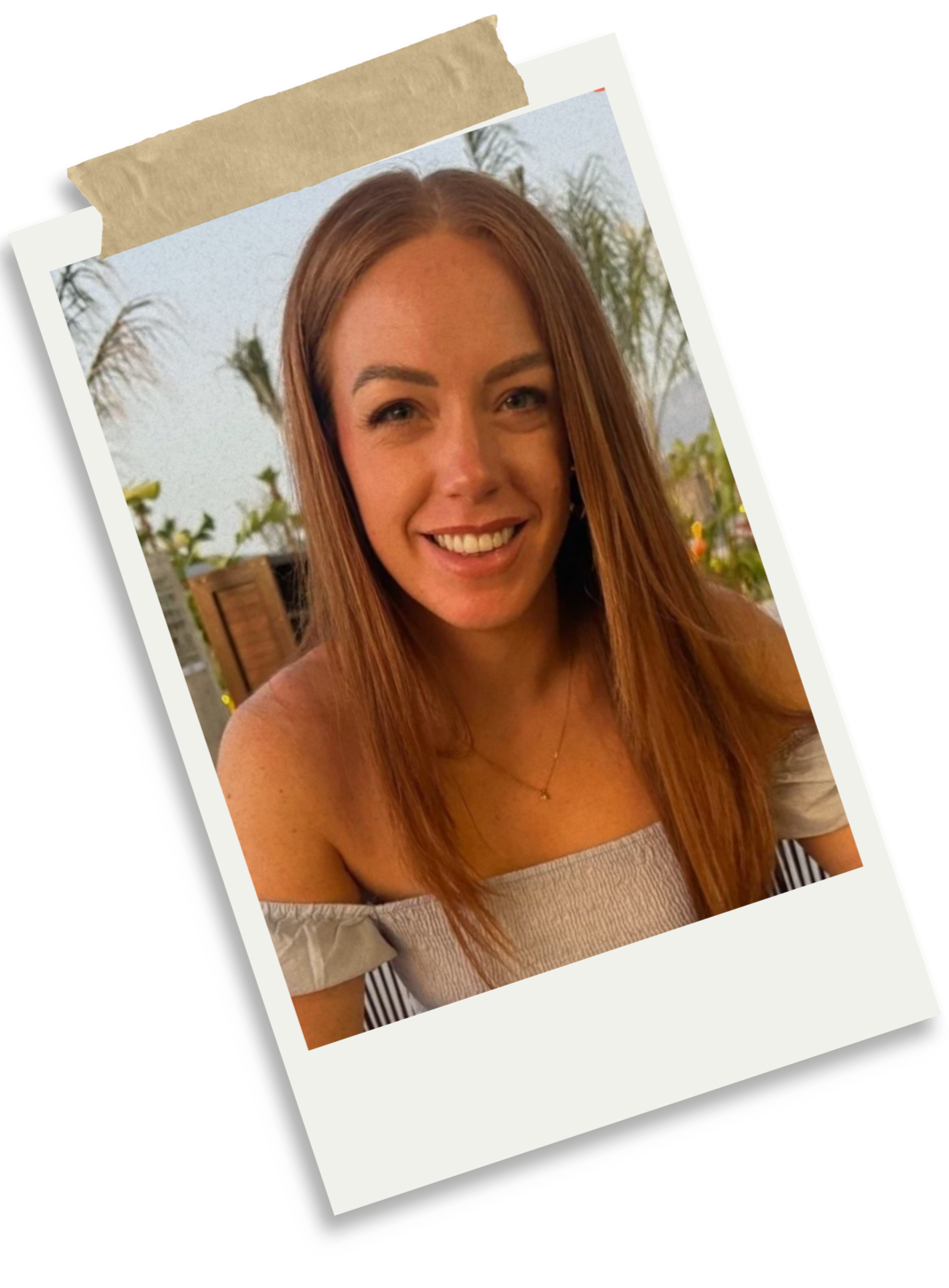 A woman with long straight red hair smiling at the camera, wearing a beige off-shoulder top and a delicate necklace, in an outdoor setting with plants and a light sky background.