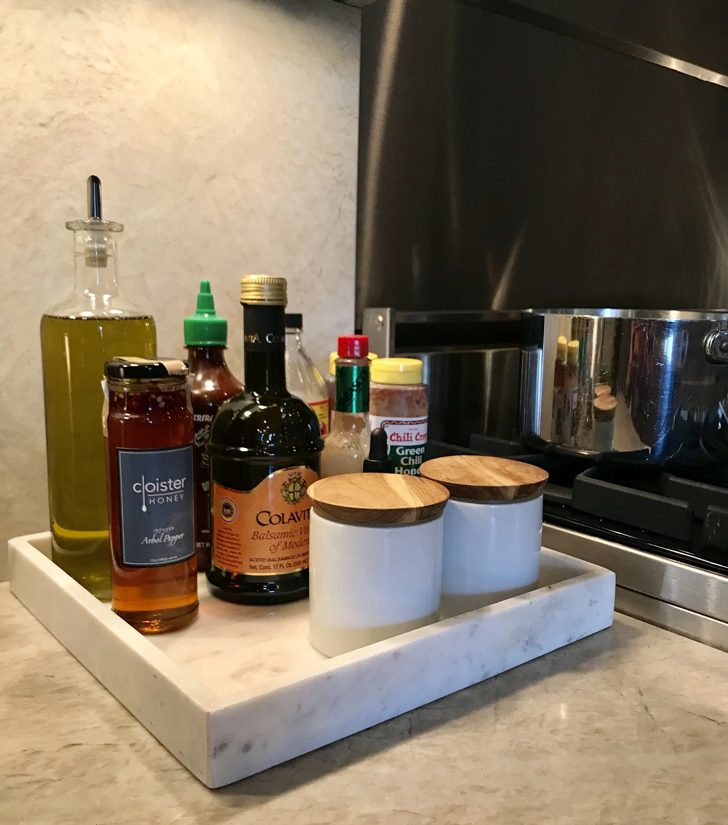 White marble countertop tray arranged with olive oil, Cloister Honey, Colavita balsamic vinegar, and two ceramic salt and pepper canisters next to a stove.