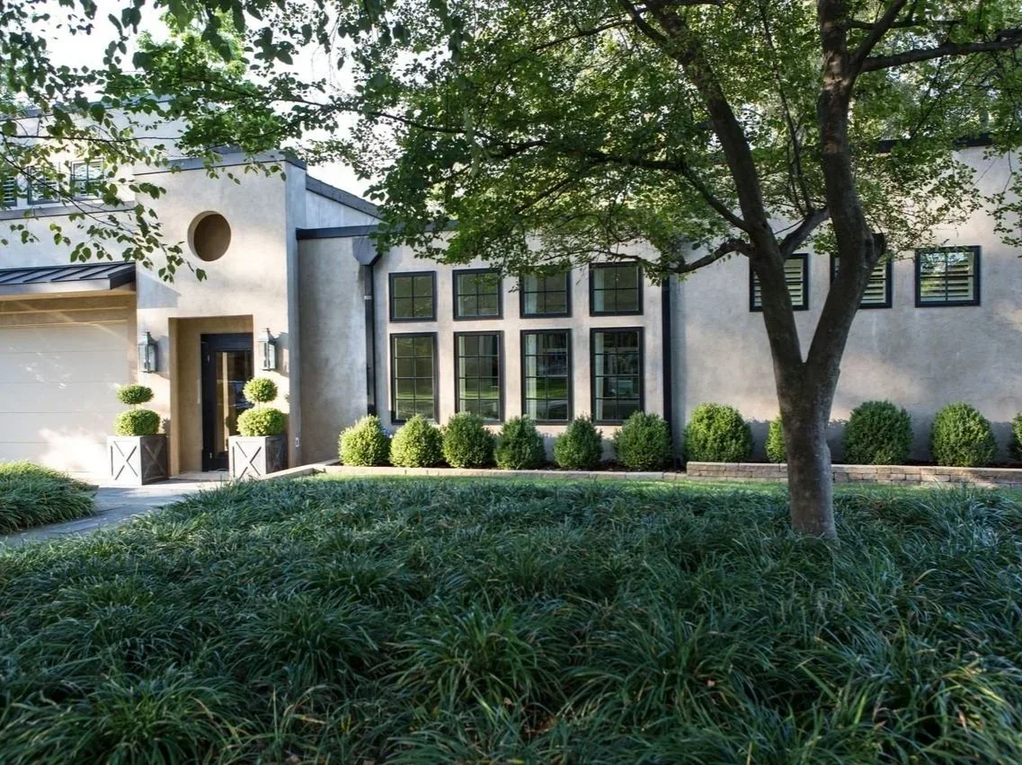 Exterior view of a modern stucco home featuring large black-framed windows, a glass front door, and manicured green landscaping with potted topiary trees.