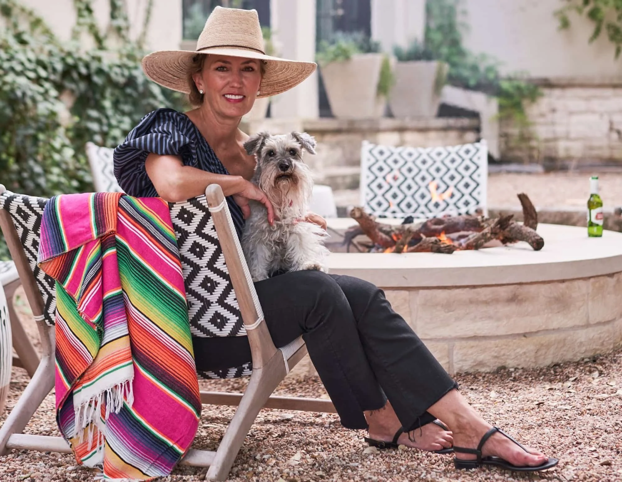 Shazalynn Cavin-Winfrey wearing a straw hat while sitting in a patio chair holding a small dog near an outdoor fire pit.