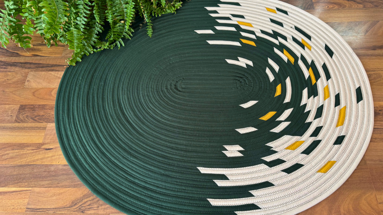 Circular Jelly Roll Rug in dark green and white with yellow accents on a wooden floor, partially covered by green fern plant.