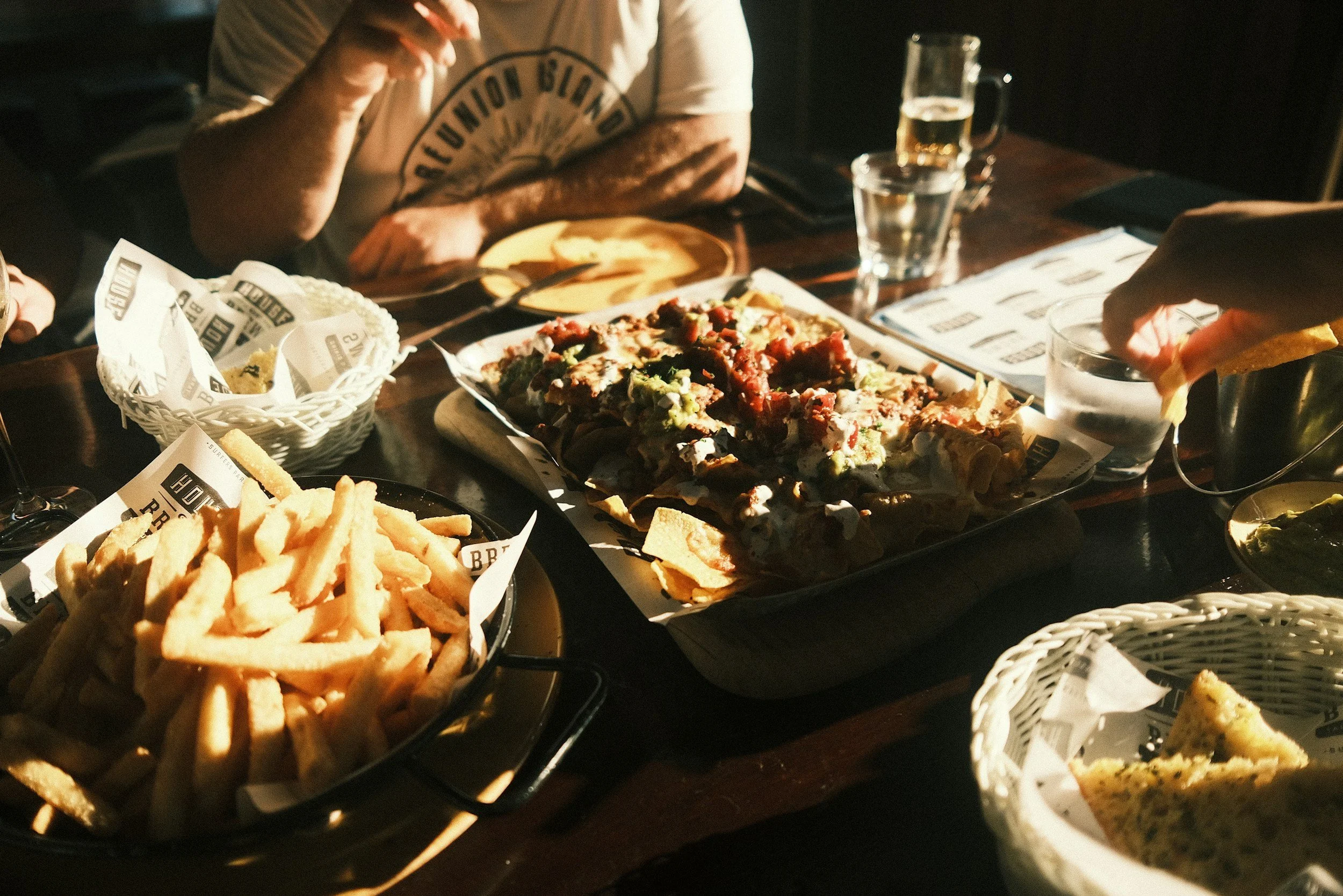 A table full of assorted dishes including fries, pizza, and bread, with drinks and people eating.