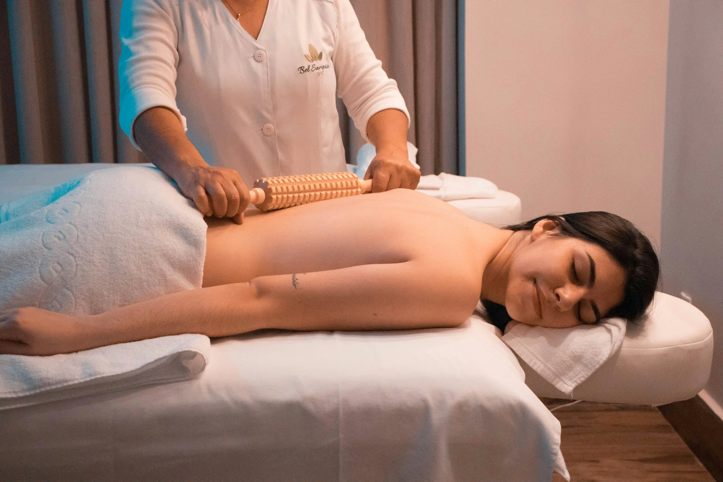 A woman is lying face down on a massage table while a massage therapist uses a wooden massage tool on her back. The woman has dark hair and is smiling with her eyes closed. The massage therapist is wearing a white uniform with a logo and is holding the tool with both hands. The room has soft lighting and neutral-colored walls.