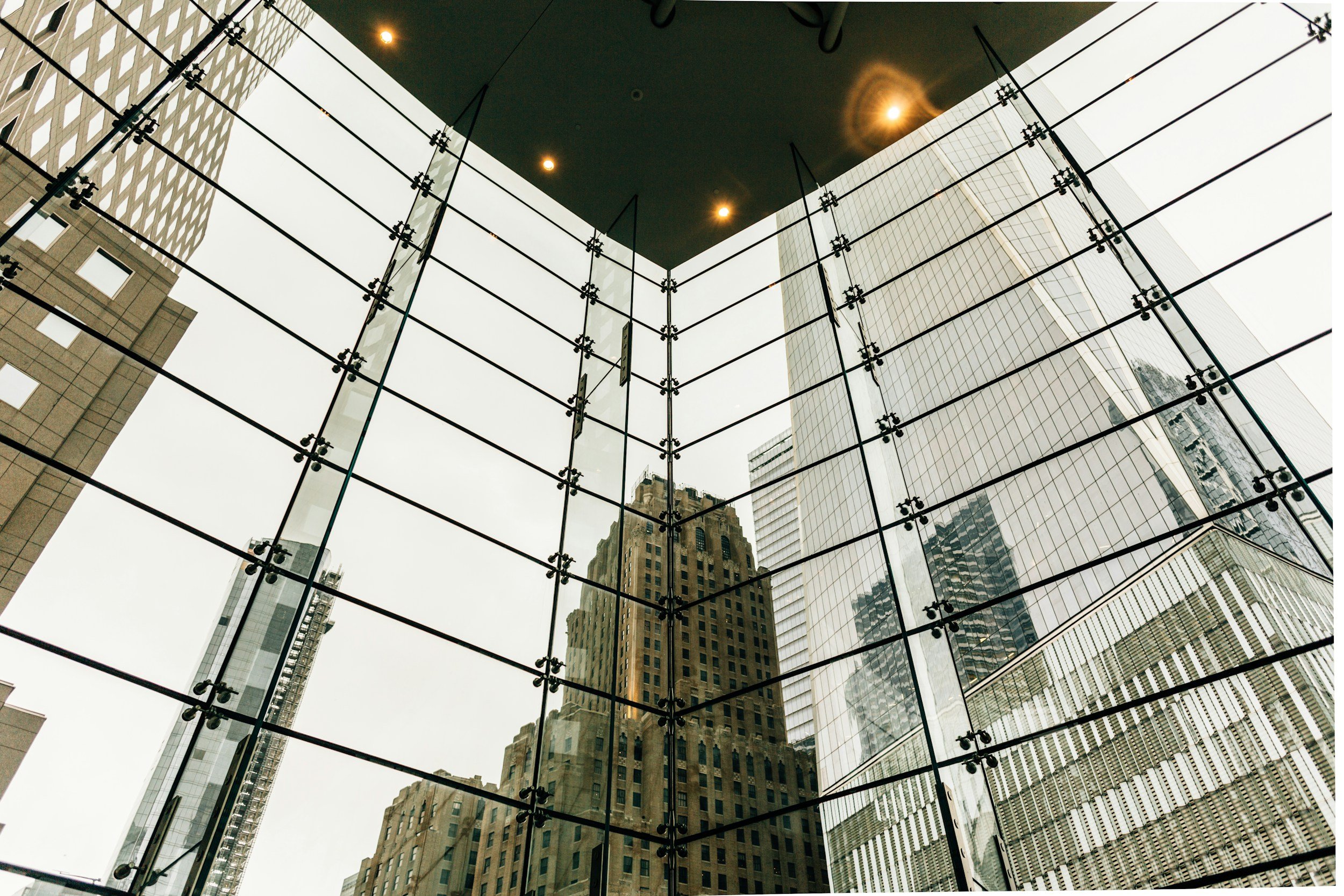 View looking up at a modern glass building with reflections of surrounding skyscrapers and a dark ceiling with ceiling lights inside the building.