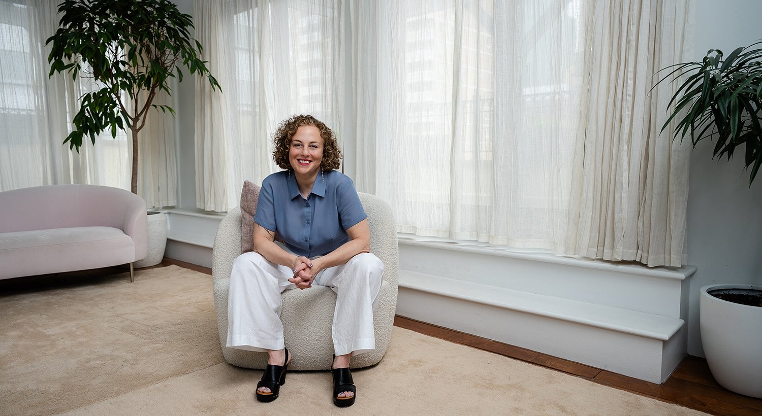 A woman sitting on a light-colored armchair in a bright room with large windows and sheer curtains, smiling at the camera.