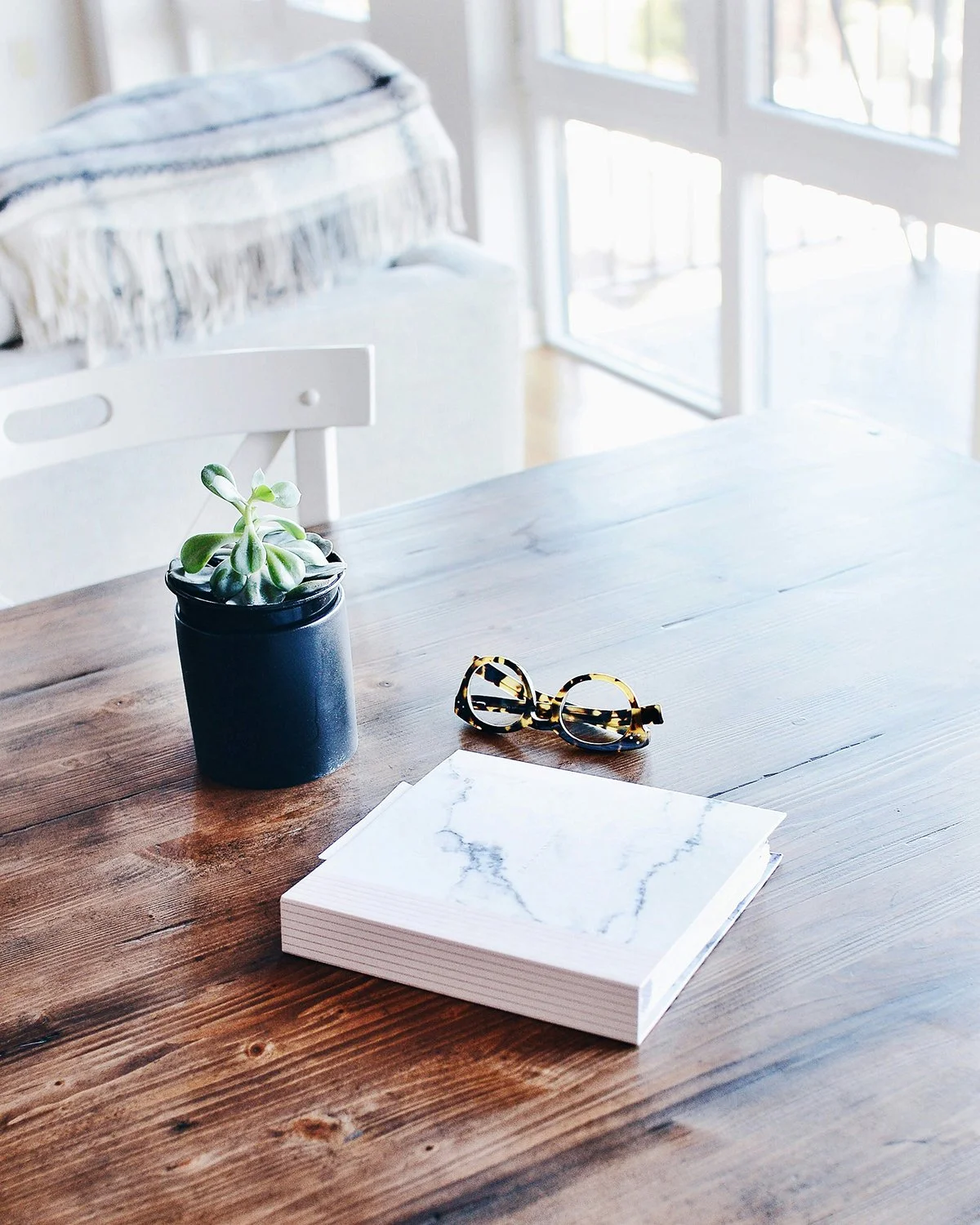 A wooden table with a potted succulent plant, a pair of tortoiseshell sunglasses, and a white marble-patterned notebook, near a window with natural light.