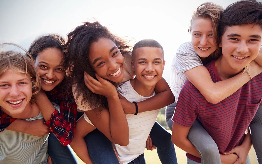 Group of smiling teenagers outdoors, some giving piggyback rides and embracing, enjoying sunny weather.