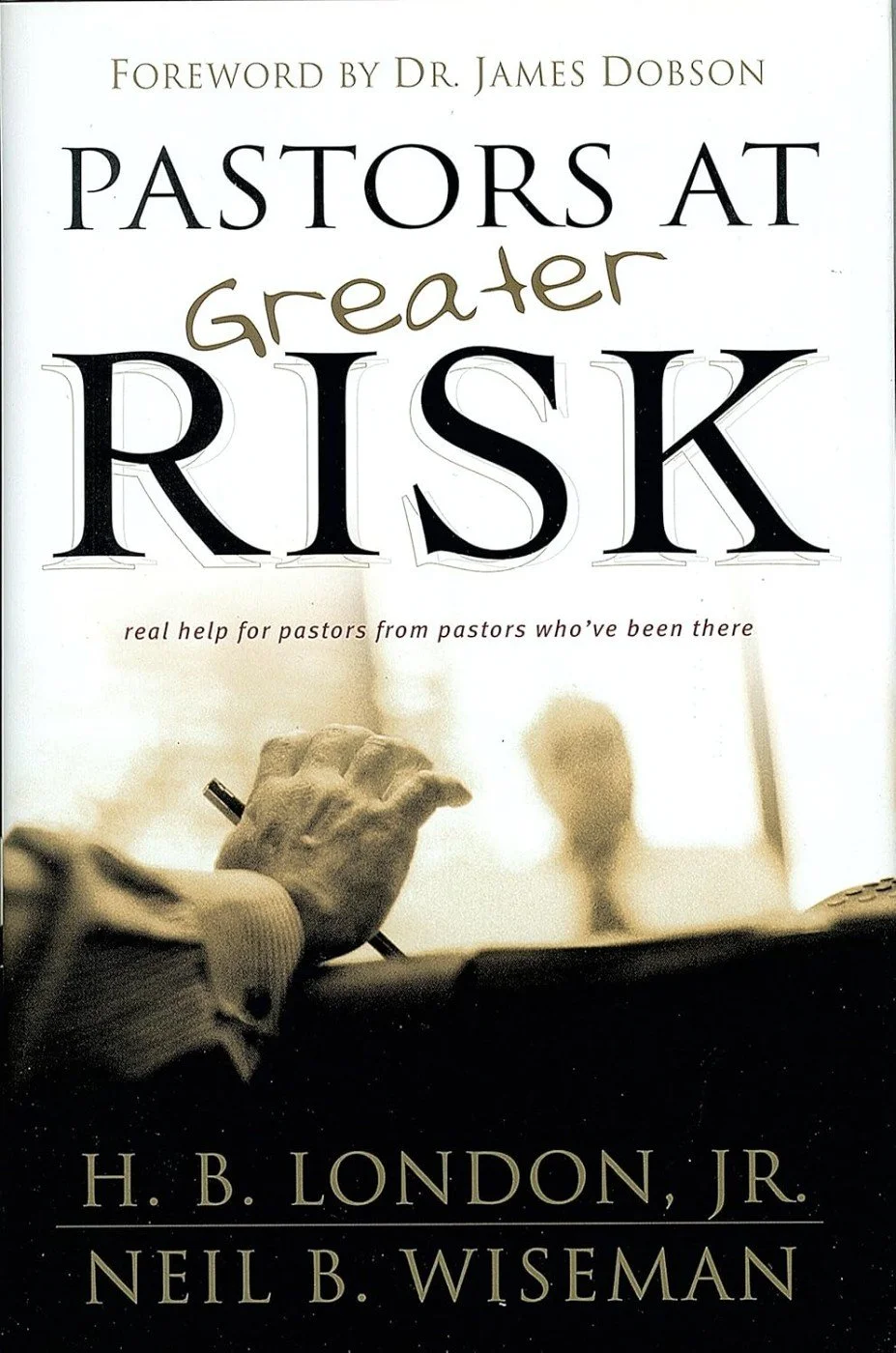 Book cover titled 'Pastors at Greater Risk' by H. B. London Jr. and Neil B. Wiseman, with a subtitle 'real help for pastors from pastors who've been there.' The cover shows a sepia-toned image of a hand holding a pen, blurred background of a person, possibly a pastor, sitting at a desk.