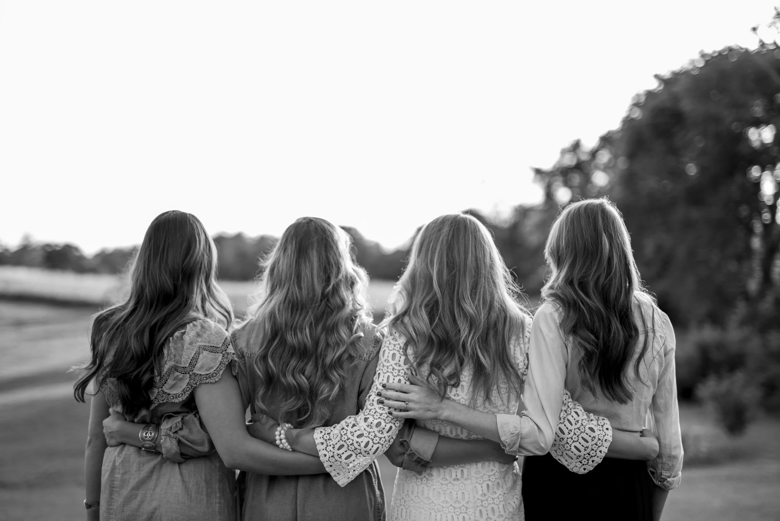Black and white photo of a group of women standing outdoors with their arms around each other, facing away from the camera, in a natural setting with trees in the background.