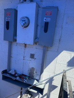 Electrical meters and panels mounted on a wall with tools on a shelf below.