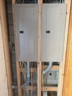 Two electrical panels mounted on a wooden wall, with conduit wiring running below.
