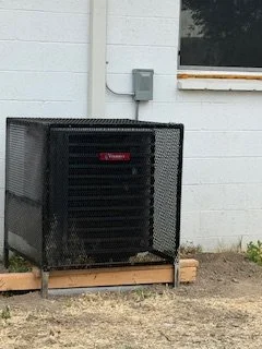 Black outdoor air conditioning unit with a protective metal cage, installed on a wooden platform outside a white building.