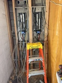 Open electrical panel with wires and breakers, and a small orange step ladder inside a unfinished wall space.