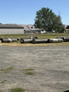 A row of large metal pipes lying on grassy ground in an open area with buildings and trees in the background under a clear blue sky.