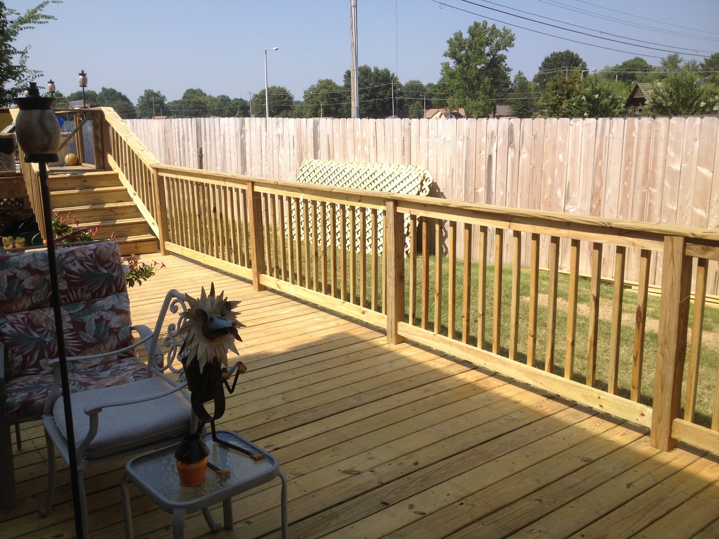 A backyard wooden deck with porch furniture, including chairs and potted plants, enclosed by a wooden fence, with trees and power lines visible in the background.