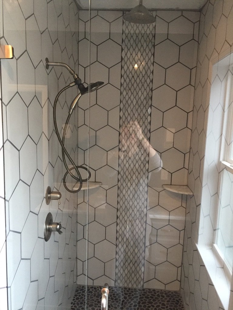 Shower with hexagonal tiles, handheld showerhead, a built-in corner shelf, and a glass shower door in a bathroom.