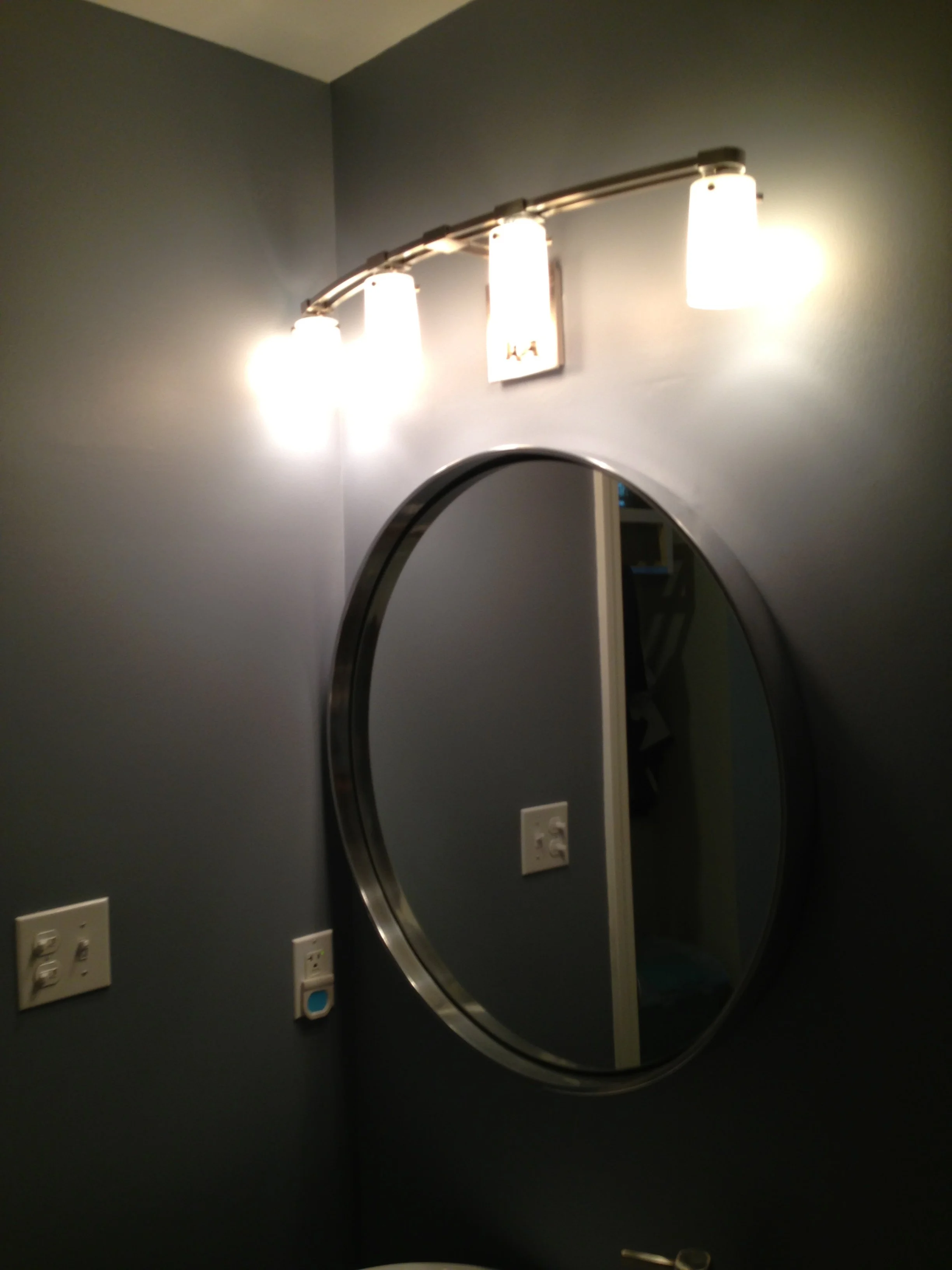 A bathroom mirror with a dark gray wall behind it, and a modern three-light vanity fixture mounted above the mirror. There is a visible outlet and a switch on the wall below the mirror.