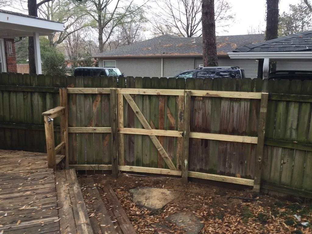Backyard with a new wooden gate installed in a weathered wooden fence; a small wooden deck with fallen leaves in the foreground, neighboring houses, trees, and parked vehicles in the background.