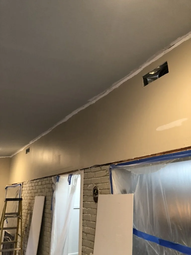 Interior of a room under renovation with a brick wall, plastic covering windows, and freshly painted ceiling and wall.