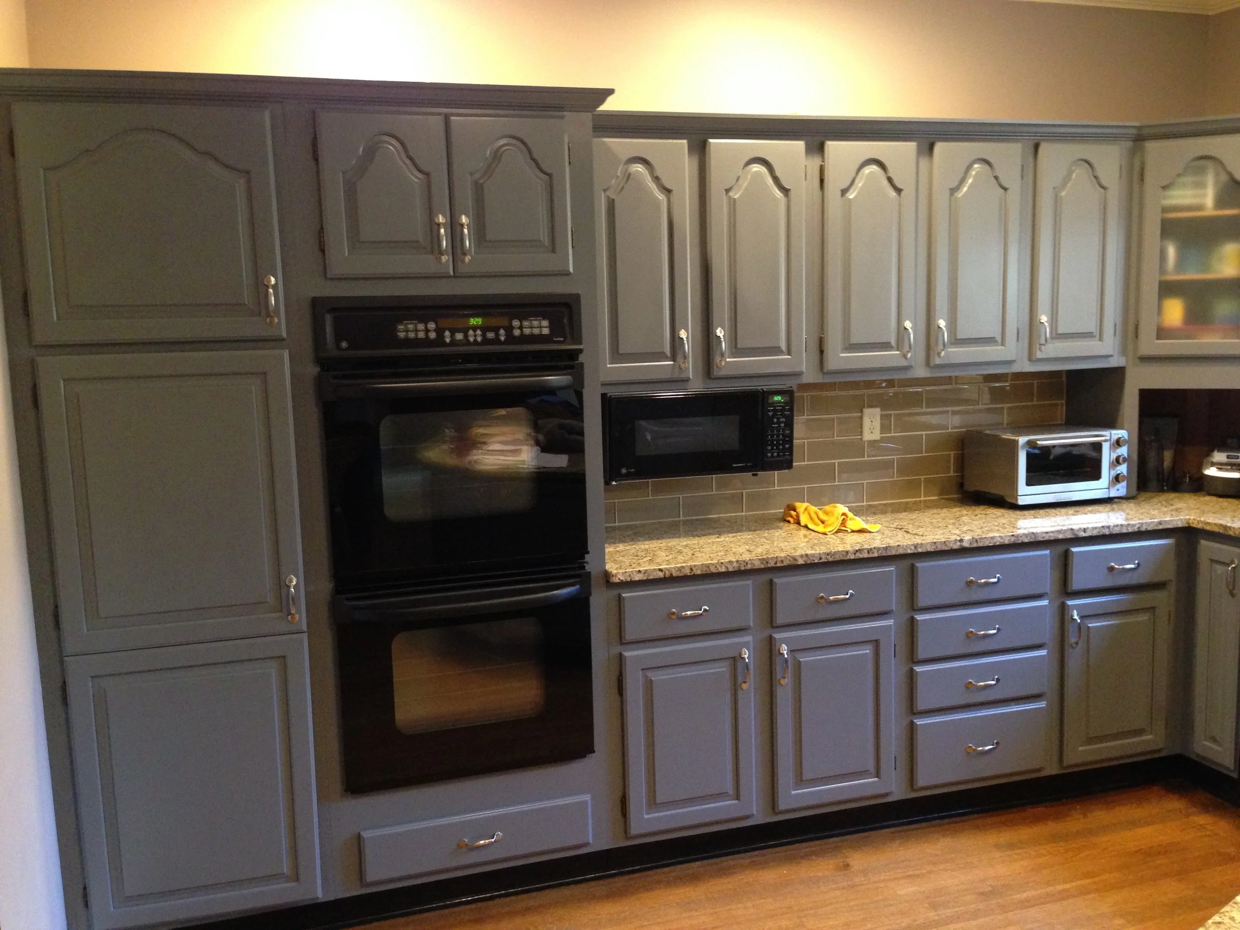 Kitchen with gray cabinets, black oven, microwave, toaster, beige wall, granite countertop, and yellow cloth on the counter.