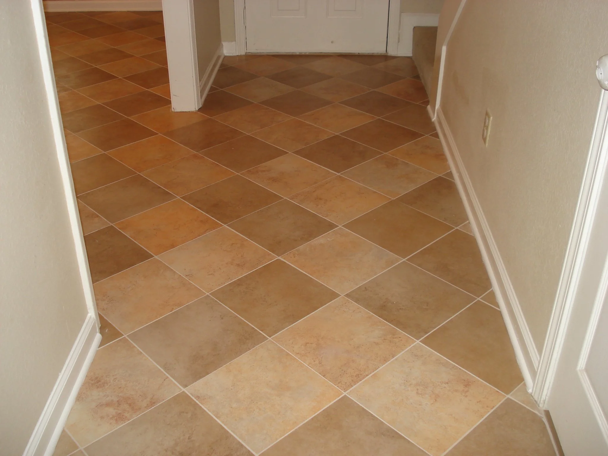 A view of a tiled floor in a hallway with a white door at the end and beige walls with white baseboards.