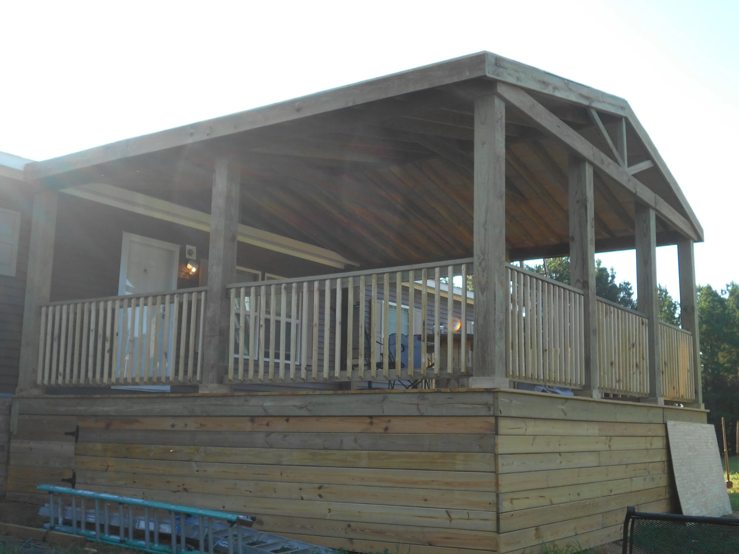 A two-story wooden deck under construction with a roof, supported by wooden beams and surrounded by a railing, attached to a house with a window and a door visible.
