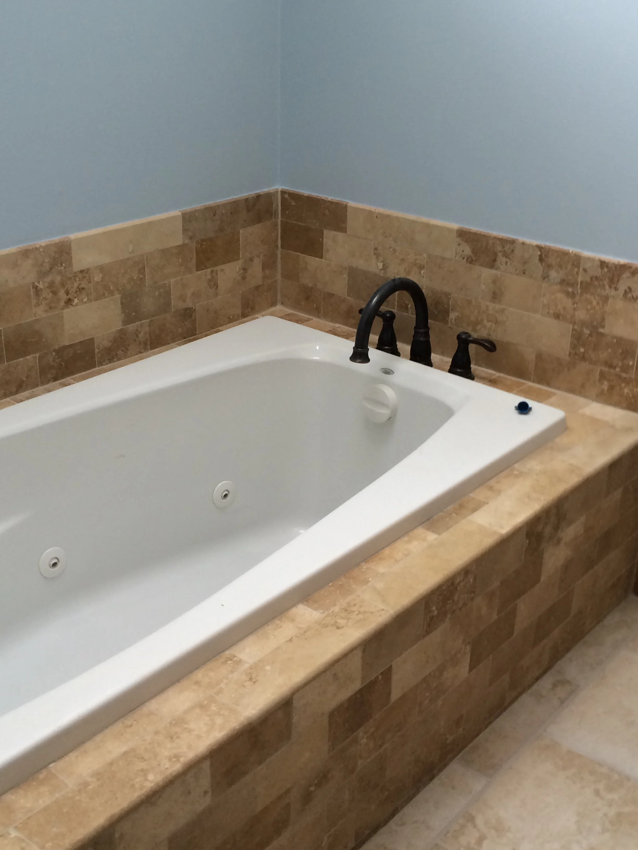 A corner bathtub with beige stone tile surround and black fixtures, located in a bathroom with light blue walls.