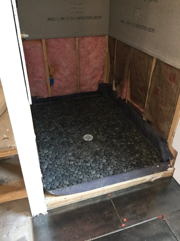A bathroom shower with a pebble stone floor, unfinished walls, and plumbing fixtures visible.