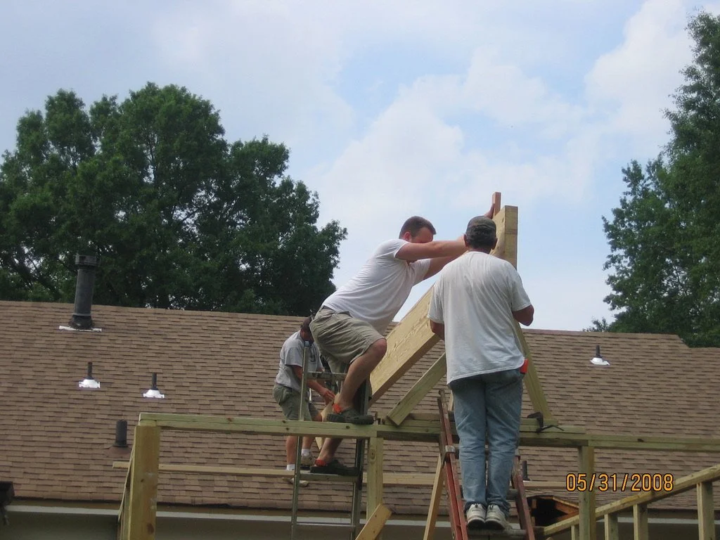 People working together on a wooden roof structure, with trees and a house roof in background.
