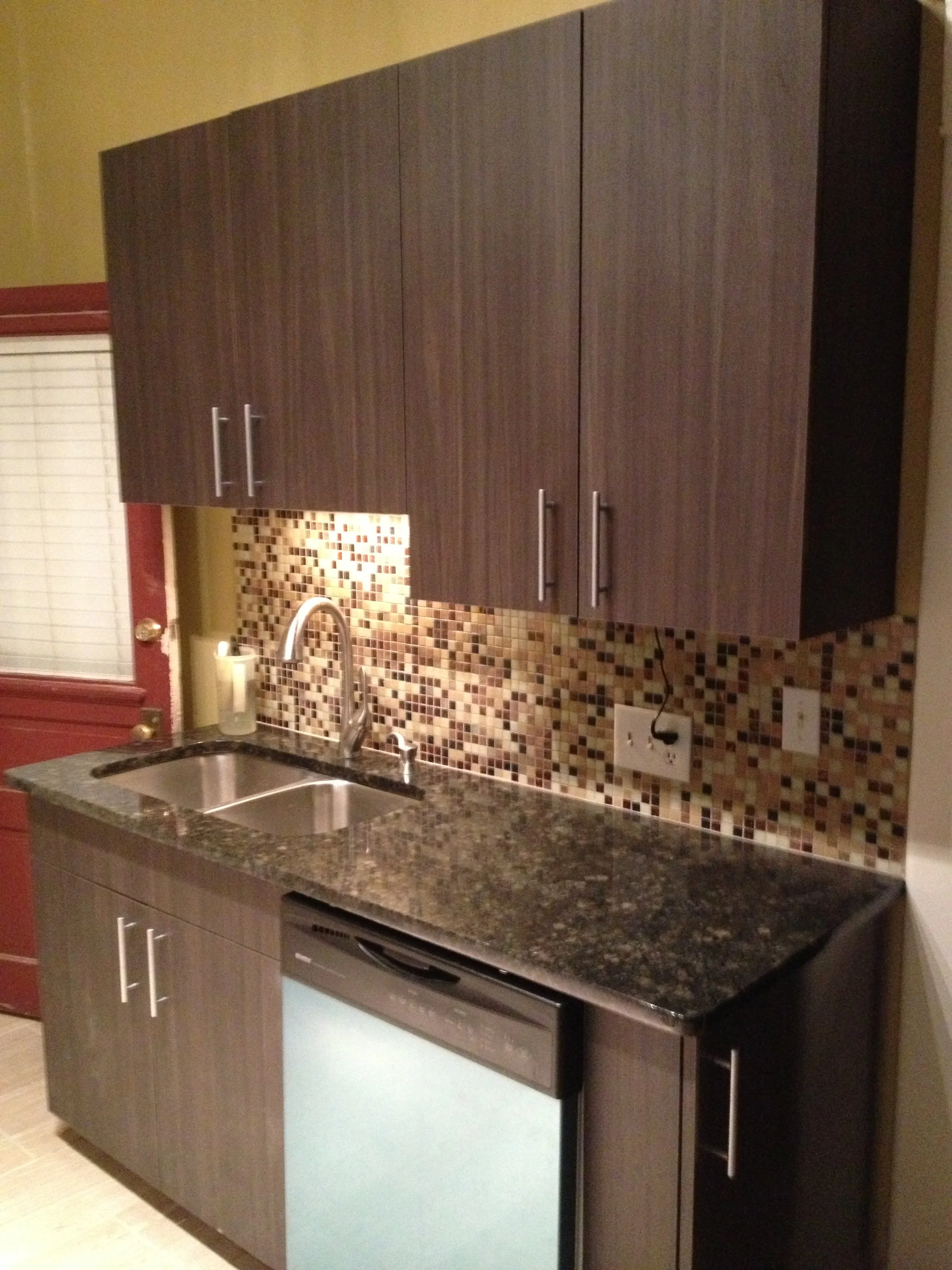 Kitchen with dark wood cabinets, mosaic tile backsplash, granite countertop, stainless steel sink, and a black dishwasher.