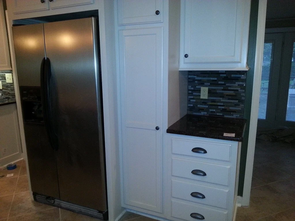 Stainless steel side-by-side refrigerator next to white kitchen cabinets and drawers with black hardware, black countertop, and a multicolored tile backsplash.