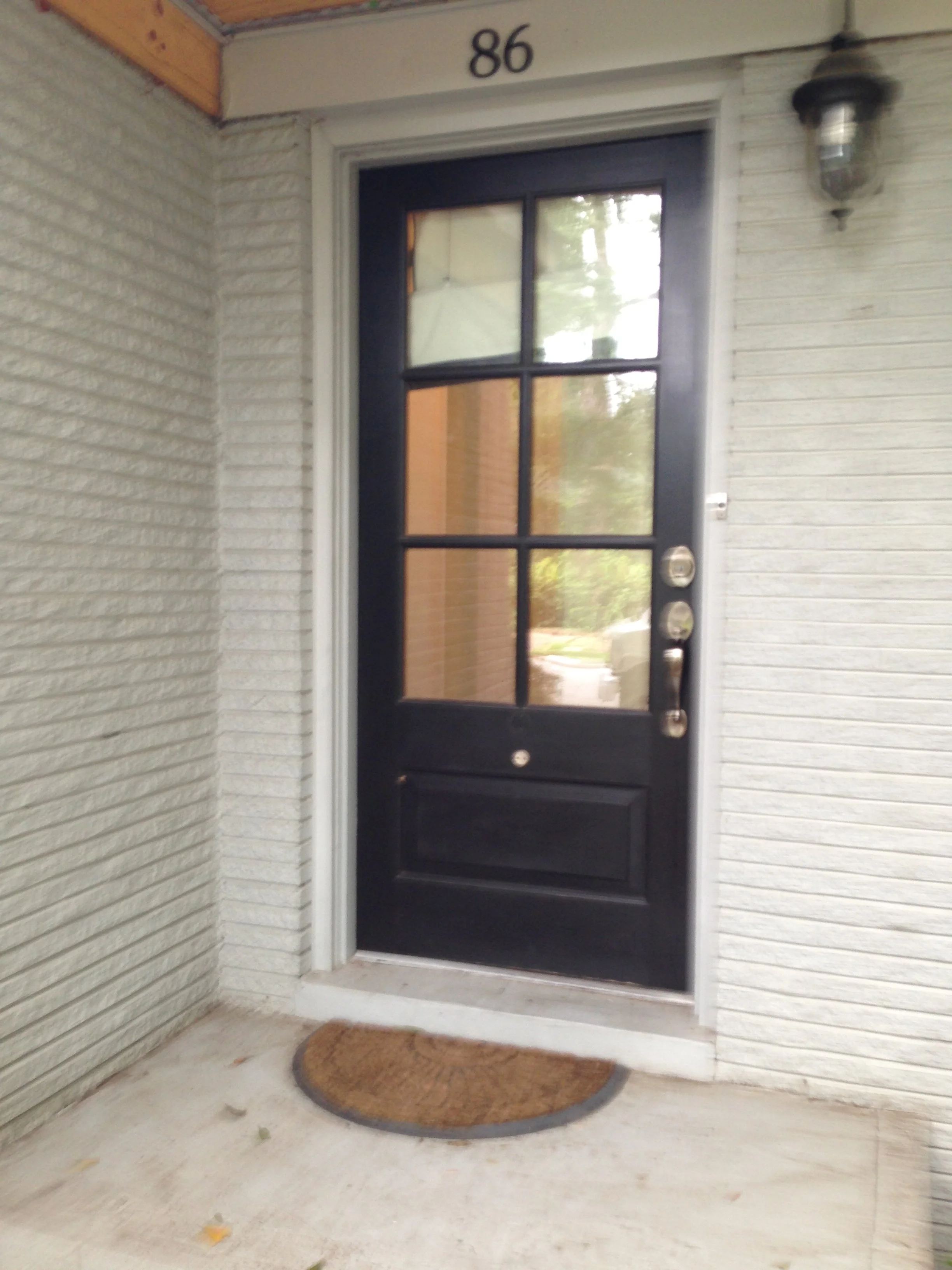 Black front door with a window pane divided into six sections, a silver doorknob, and a deadbolt lock. A brown doormat is in front of the door. The house number '86' is above the door, walls are white with textured paneling on the right and horizonta