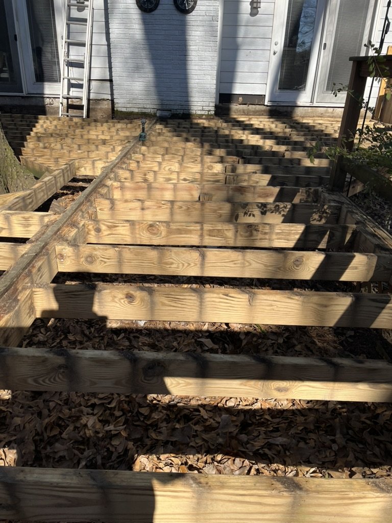 Wooden deck framing under construction outside a house, with stairs leading up to a back door; fallen leaves on the ground.