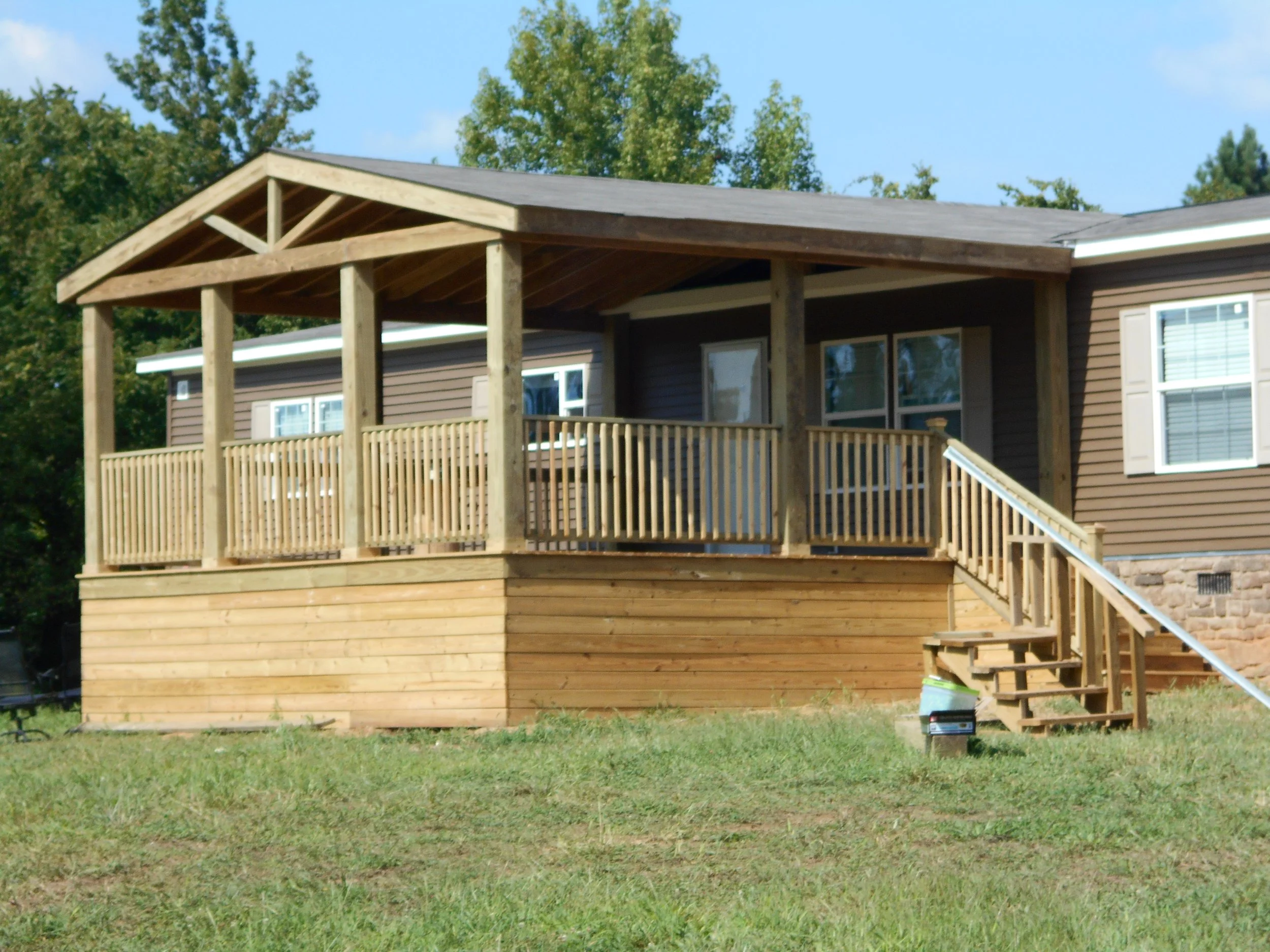 A house with brown siding and white-framed windows, surrounded by trees, with a newly built wooden deck and stairs leading up to it, in a grassy yard under a blue sky.