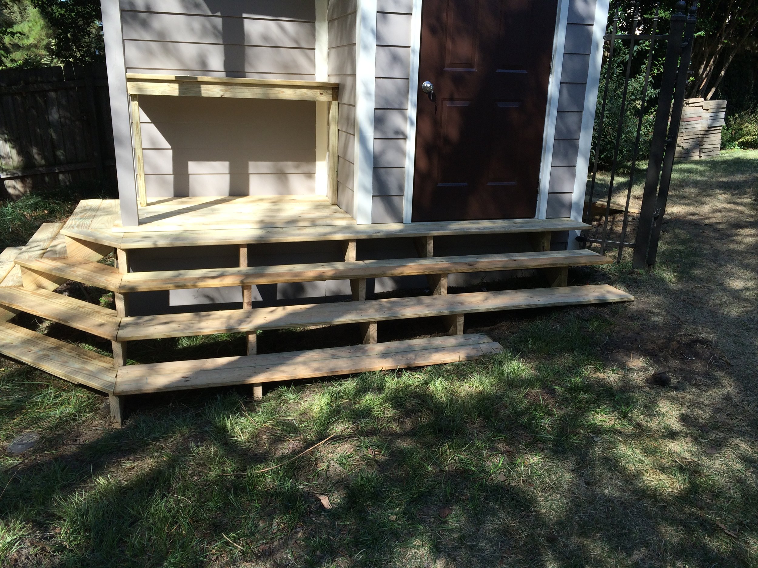 A small outdoor shed or playhouse with wooden stairs, a brown door with a doorknob, and a partially constructed deck with new wooden steps. The shed has light-colored horizontal siding, and the area is surrounded by grass, trees, and a black metal fe
