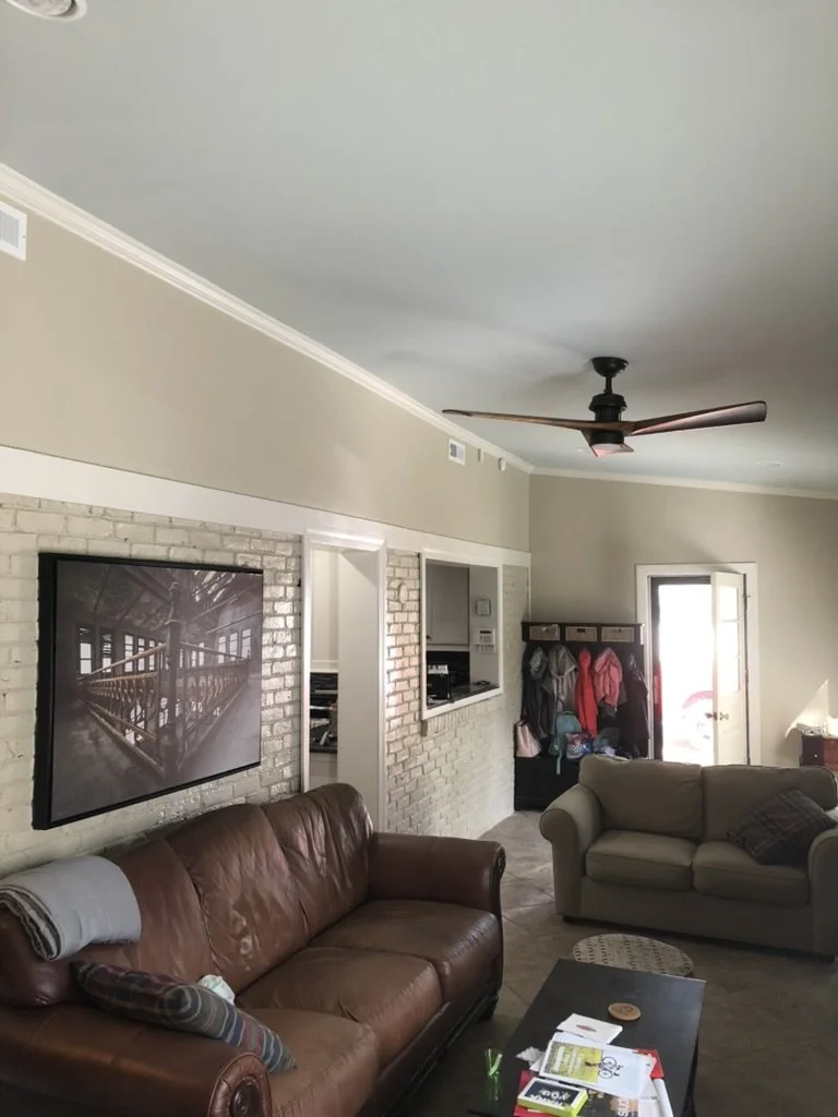 Living room with leather and fabric couches, wall-mounted TV, brick accent wall, ceiling fan, coat rack near door, and a coffee table with papers and a coaster.