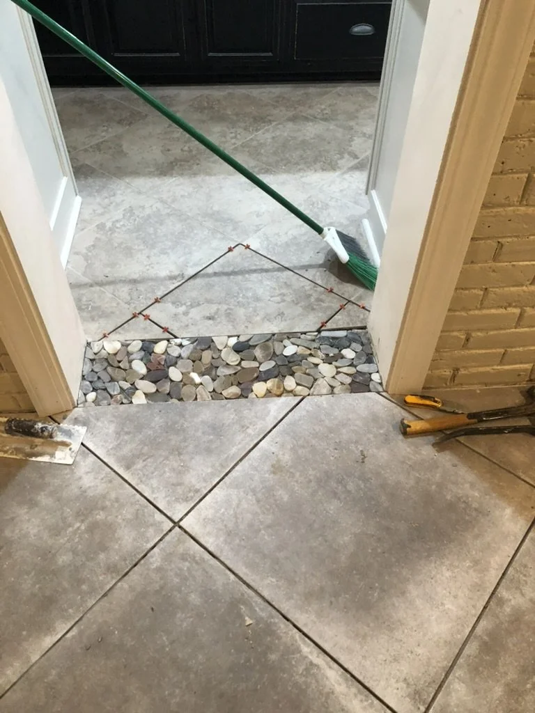 This is a photo of a home renovation or flooring project showing installed large gray ceramic tiles and an area of small river rocks. There are tools such as a trowel and a tile cutter on the floor, along with a broom leaning against the wall. A chal