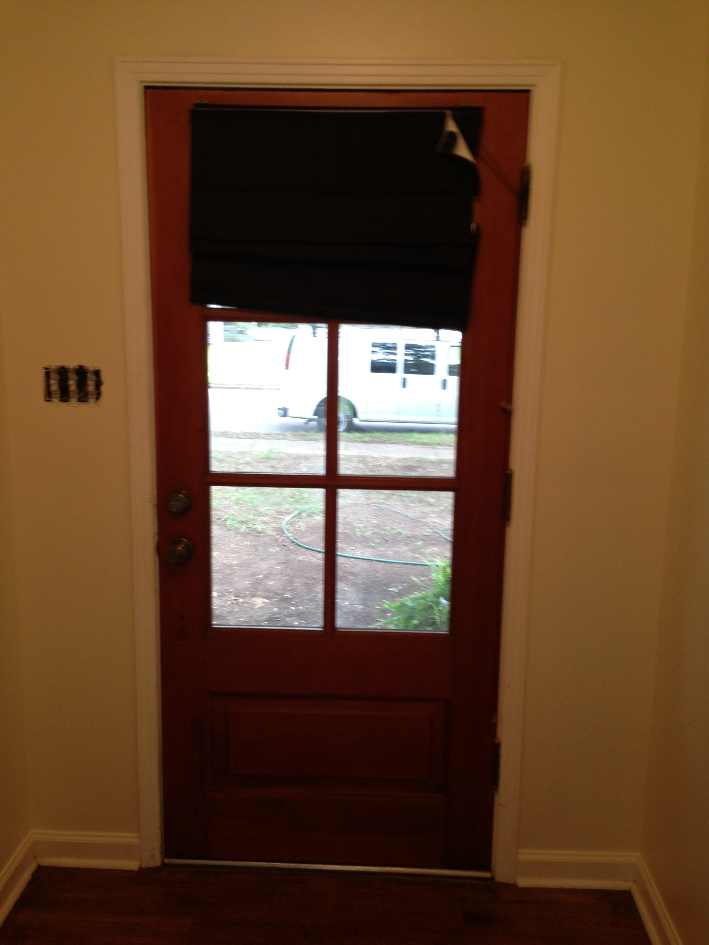 A wooden door with a glass window, partially covered by a closed black blackout curtain, with a view of the outside street and a white vehicle parked nearby.
